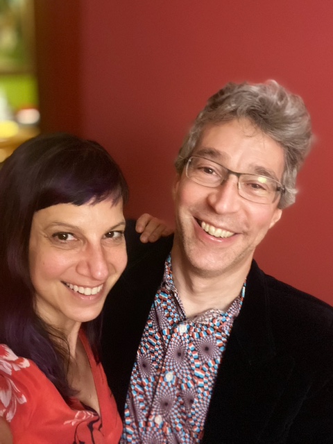 Wendy and Jon are embracing and smiling at the camera in front of a red background