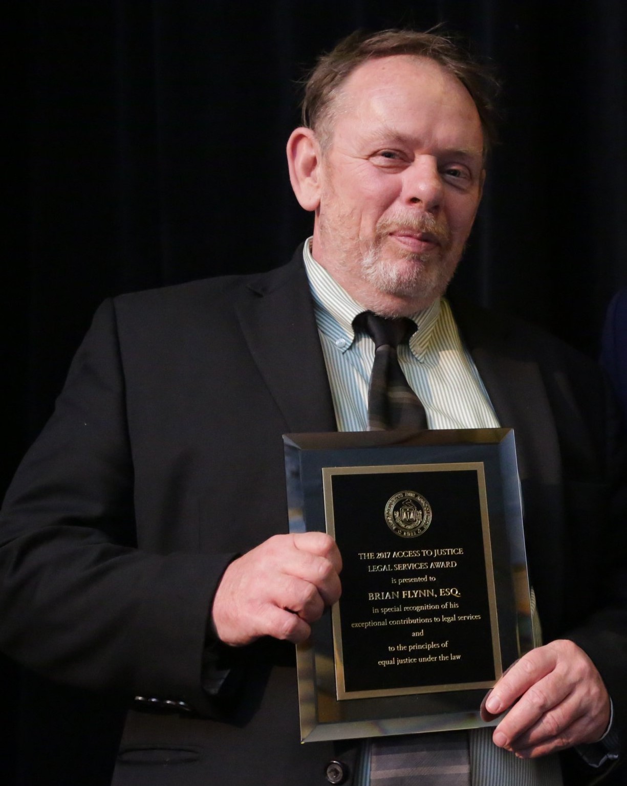 Brian accepts an award from the Mass Bar Foundation. He is standing on stage holding an award plaque and wearing a dark suit.
