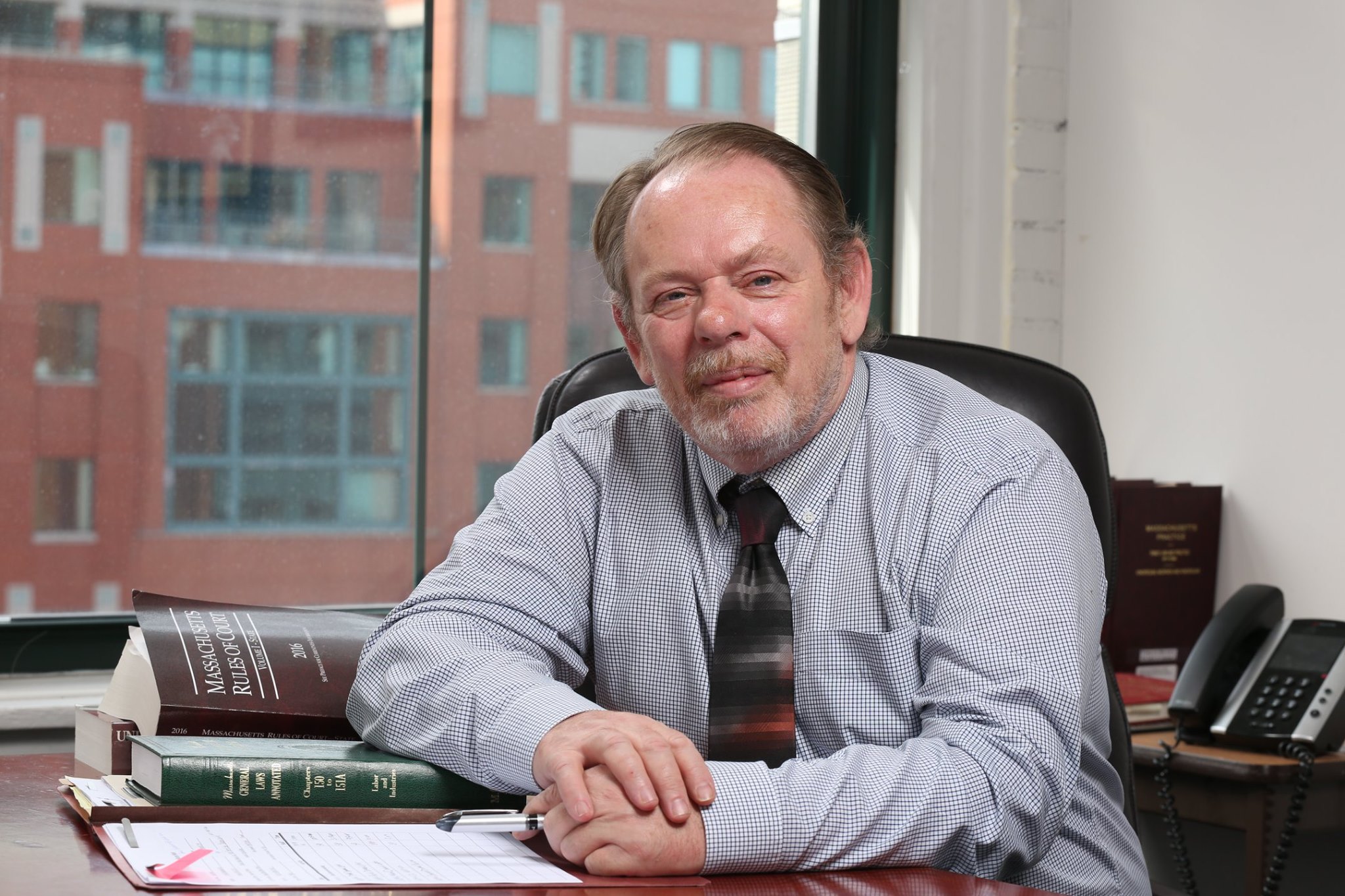 Older photo of Brian wearing a suit and sitting at a conference table with a stack of legal books and documents
