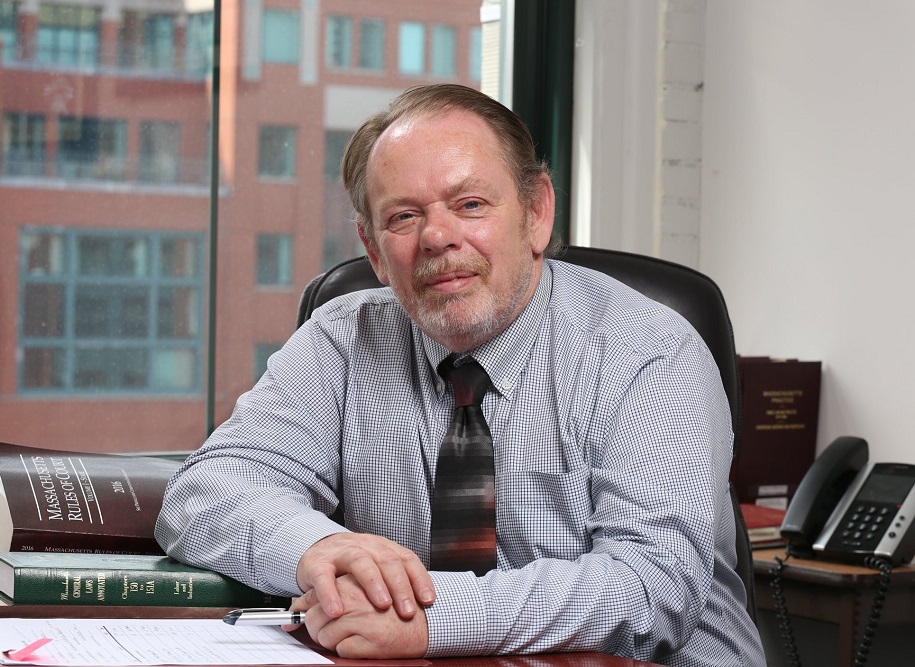 Photo of Brian Flynn sitting in a GBLS office with files and law books on the table in front of him. Brian is wearing a button down shirt and a tie.