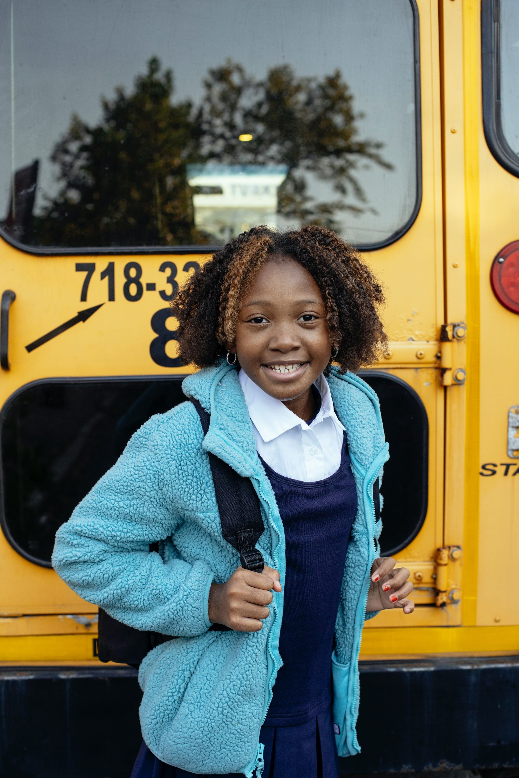 Photo of girl carrying a backpack, standing in front of a school bus. 