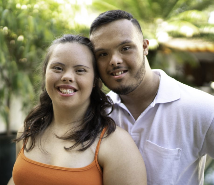 Photo of two people: a young man with disabilities wearing a white button down shirt, and a young woman with disabilities wearing an orange tank top. They are standing outdoors, close together, and smiling at the camera.