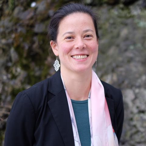 Liliana Ibara stands smiling at the camera, she is wearing a teal blouse, a pale floral scarf, and a navy blazer. She is outside in front of some rocks.