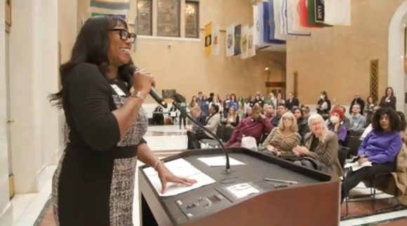 Still image from GBLS' video, features a woman speaking into a microphone in front of an audience