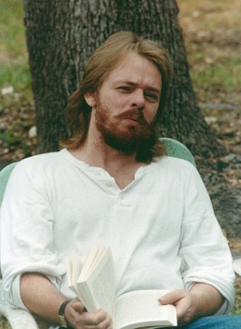 A picture of Brian Flynn in his 20s. Brian has long hair and a beard, he is wearing a white shirt and is sitting down outdoors holding a book.