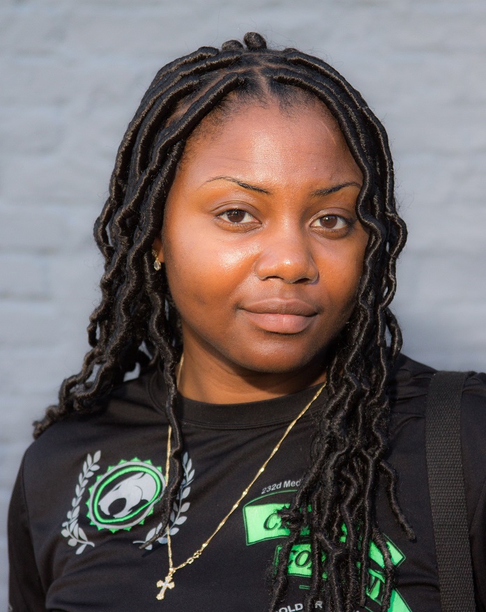 Photograph of a 20-something African American woman looking at the camera. She has a slight smile and is wearing a black shirt and has long hair.