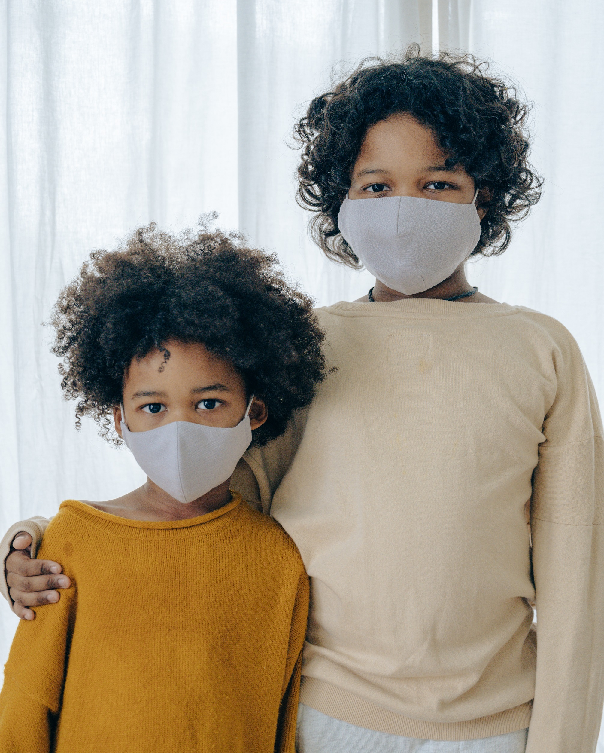 Photo of two siblings standing side by side, dressed in pastel colors and wearing face masks.