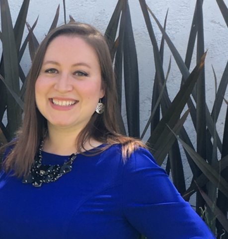 Ana Cruz, Chief Development Officer, is wearing a blue dress and black necklace and smiling in front of some tall plants.