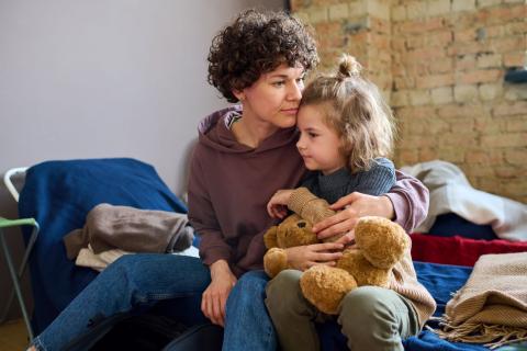 woman sitting on a cot holding a child with a teddy bear in her lap