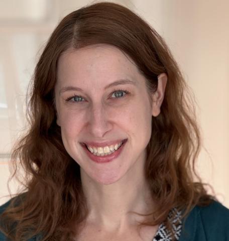 Ilana Gelfman, is a white woman with brown hair. She is smiling at the camera in front of a neutral background.