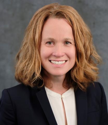 Zoe Cronin, Deputy Director, is a white woman with auburn hair. She is wearing a white blouse and black blazer and is smiling at the camera in front of a neutral background.