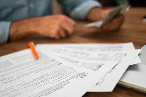 Close-up of tax documents and forms with a pen on a desk, while a person in the background counts cash